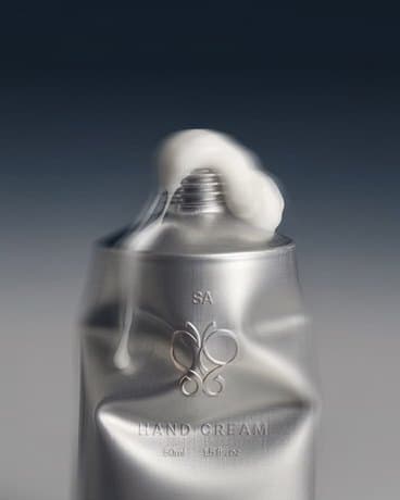 A silver embossed hand cream tube photographed against a graduated grey background, cream at the nozzle