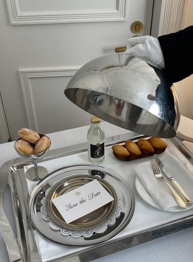A gloved hand lifting a silver cloche from a room-service tray set with madeleines and a Save the Date card