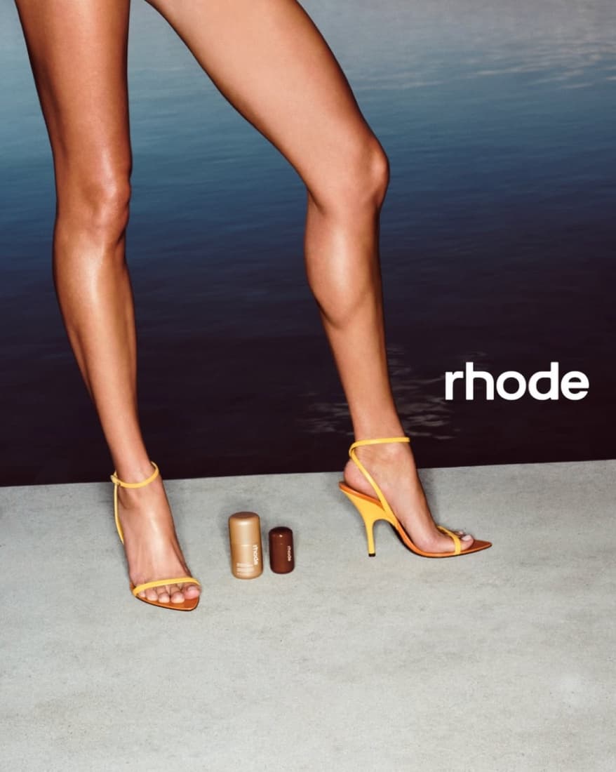 rhode campaign photograph — bare legs in strappy yellow heels beside two lip products on a concrete ledge above water