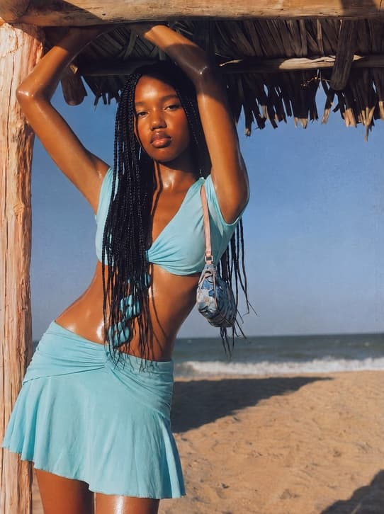 Launch campaign photograph of a model with long braids in a light blue two-piece standing on a sunlit beach